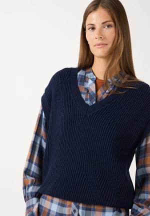 Junge Frau trägt eine marineblaue gestrickte Weste über einem blau-braun-weiß karierten Hemd und steht vor einem schlichten Hintergrund.