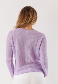 Donna con capelli castano chiaro che indossa un maglione a maglia lilla e pantaloni bianchi, mostrata di spalle su uno sfondo semplice.