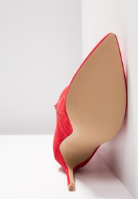 Scarpa con tacco alto in suede rosso, con punta affusolata e suola in gomma beige, caratterizzata da una superficie liscia e un tacco a stiletto sottile.