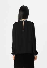 Blouse noire à manches longues avec un dos en trou de serrure, un col orné et des détails de poignets élastiques. Texture lisse avec un design fluide.