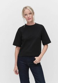Musta lyhythihainen paita, joka on valmistettu puuvillasta ja jossa on pyöreä pääntie. Paidan malli on rento ja minimalistinen, ja sitä on yhdistetty tummiin farkkuihin.