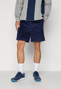 Marineblå corduroy-shorts har en avslappet passform og teksturert overflate. De er kombinert med en grå og marineblå fargeblokkerte genser og mørke treningssko.