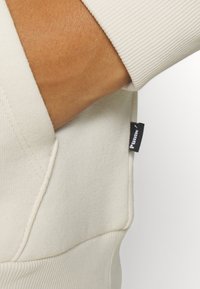 Krämfärgad sweatshirt med ribbad mudd, med en sidficka och en liten svart etikett med vit text sydd på fållen.