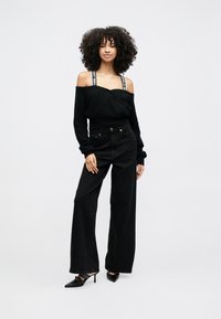 Kvinna med lockigt hår som bär svart off-shoulder tröja med märkta band, vida svarta jeans och svarta spetsiga klackar, står mot vit bakgrund.