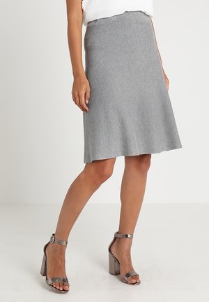 Femme portant une jupe grise longueur genoux avec une taille festonnée et des sandales métalliques à talons hauts avec bride à la cheville, debout sur un sol blanc.