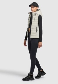 Helles graues Pufferweste mit Kapuze, mit Reißverschlusstaschen, kombiniert mit einem schwarzen Langarmoberteil und Leggings sowie schwarzen Sneakers. Glatte Textur.