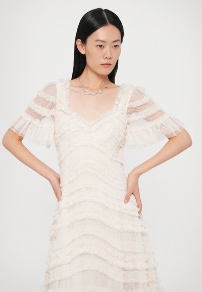 Λευκό φόρεμα με στρωματωτές βολάν, διαφανείς μανίκια και V-λαιμόκοψη. Το μοντέλο έχει διακοσμήσει με ασημένιο κολιέ. Ομαλό ύφασμα με υφή.