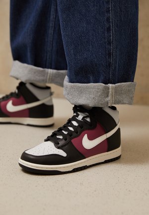 Personne portant un jean bleu retroussé et des baskets montantes Nike noires, blanches et bordeaux sur un sol clair.