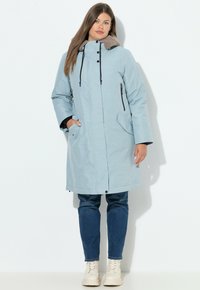 Giacca lunga azzurro chiaro con cappuccio foderato in beige morbido, dettagli con zip nera, tasche a toppa e vestibilità ampia. Abbinata a jeans e stivali bianchi.