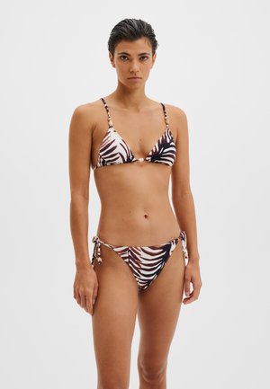 Nainen, jolla on lyhyet tummat hiukset ja päällään seeprakuosinen kolmionmuotoinen bikini, jossa helmikoristeita, seisoo yksinkertaisen vaalean taustan edessä.