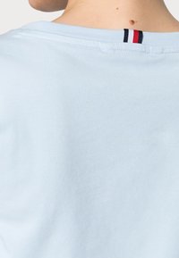 Světle modré tričko s kulatým výstřihem, na zadní straně s malou černou, červenou a bílou pruhovanou štítkem. Měkká, hladká textilie.