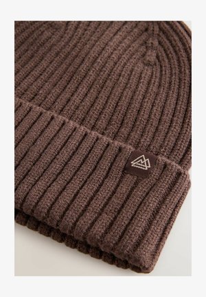 Barna kötött sapka bordás textúrával és hajtogatott szegéllyel. Kis szőtt címkével rendelkezik, amelyen hegy design található. Meleg anyagból készült.
