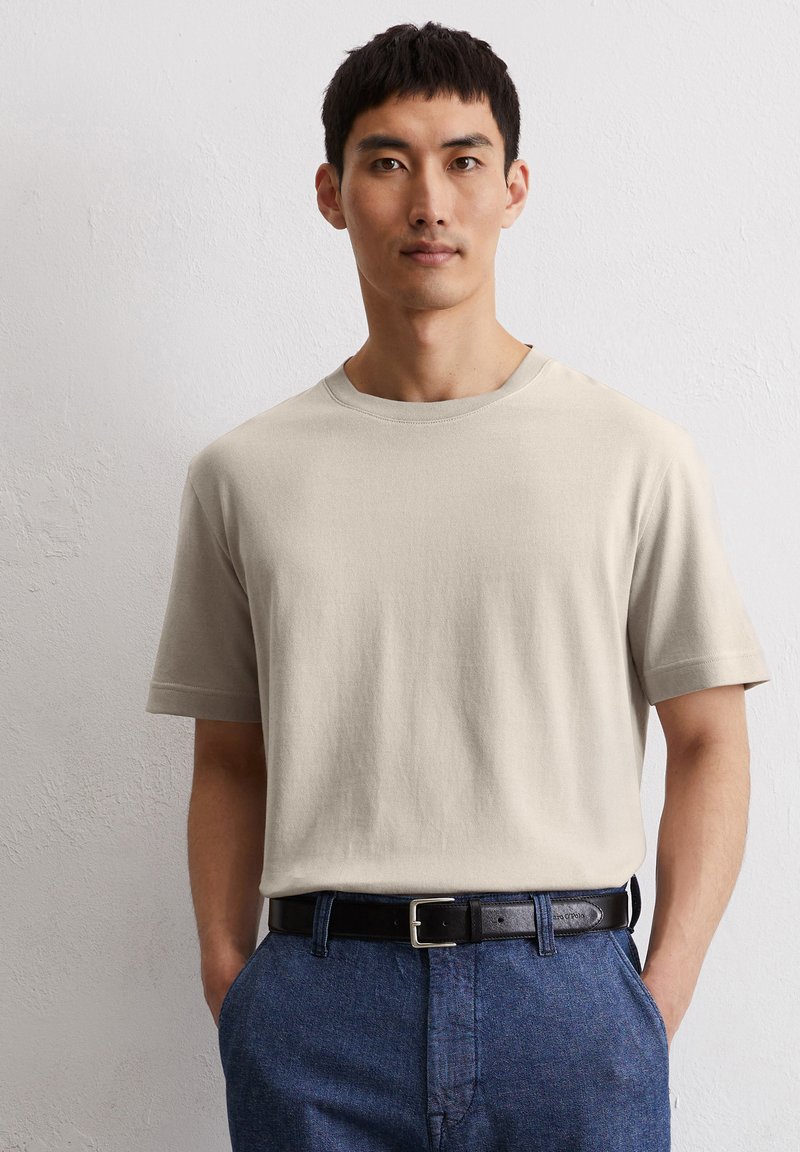 Beige Baumwoll-T-Shirt mit kurzen Ärmeln und Rundhalsausschnitt, kombiniert mit blauen strukturierten Hosen und einem schwarzen Leder-Gürtel mit einer silbernen Schnalle.