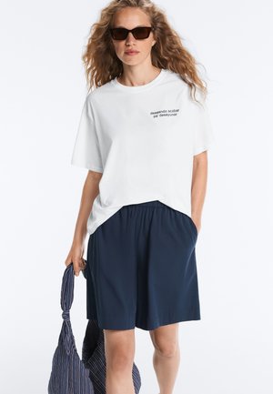 Femme portant un t-shirt blanc avec un texte en espagnol, un short bleu marine, des lunettes de soleil et tenant un sac tote rayé bleu marine avec un nœud.