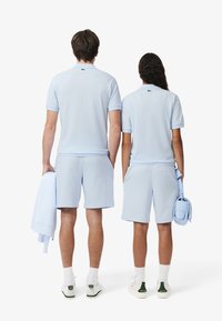 Polo shirt e pantaloni corti di un blu chiaro, realizzati in un tessuto morbido, con un logo discreto sul retro. Gli accessori includono un asciugamano e una borsa piccola.