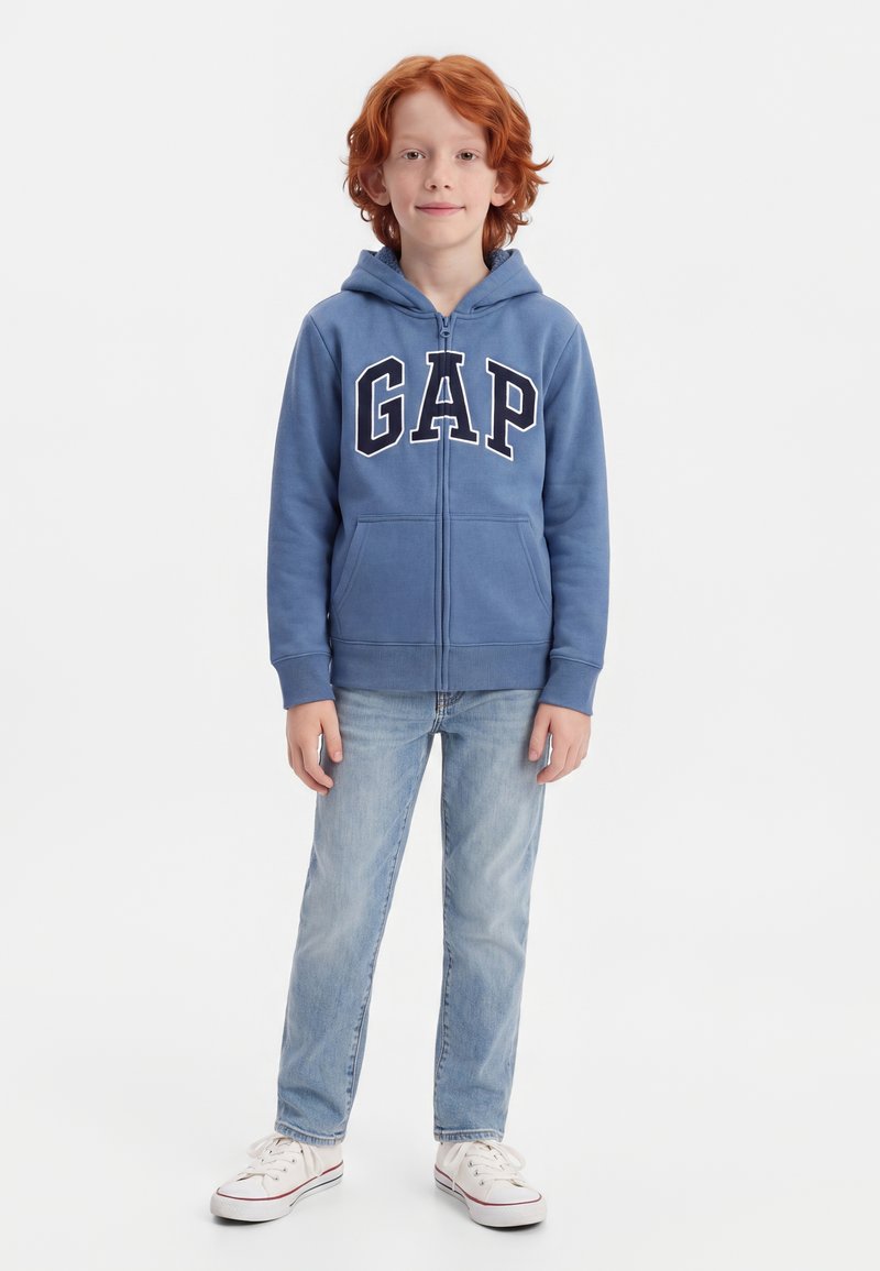 Enfant aux cheveux roux portant un sweat à capuche zippé bleu GAP, un jean bleu clair et des baskets blanches, debout sur un fond blanc.