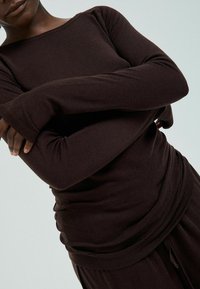 Person wearing a dark brown long-sleeve shirt and matching pants, with arms crossed over the torso against a light background.