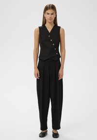 Ženska v brezrokavnem črnem gumbu in ohlapnih črnih hlačah, stoji pred enobarvno belo kuliso.