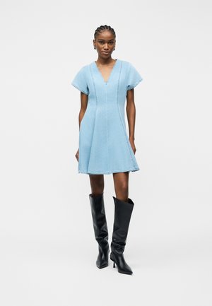 Ženska stoji oblečena v svetlo modro krilo A-linije z kratkimi rokavi in V-izrezom, obuta v črne škornje s peto do kolen, na enobarvnem ozadju.
