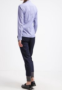 Woman wearing a light blue button-up shirt, navy jogger pants with gray cuffs, and black dress shoes standing against a white background.