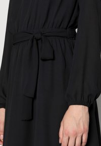 Robe noire à manches longues et ceinture nouée à la taille. Fabriquée en tissu doux avec une texture lisse et un détail froncé à la taille.