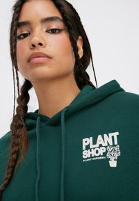 Green hoodie with "PLANT SHOP" logo and plant graphic. Features drawstrings and a soft texture, framing the face with braided hair.