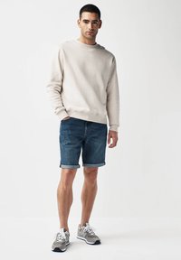Hellbeige Sweatshirt mit rundem Ausschnitt, Denim-Shorts und grauen Sportschuhen. Die Shorts haben aufgerollte Säume; das Sweatshirt hat eine glatte Textur.