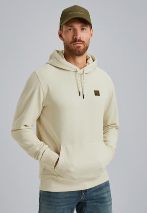 Man in beige hoodie met kleine patch en groene pet, staande met handen in de hoodie zakken, neutrale achtergrond.