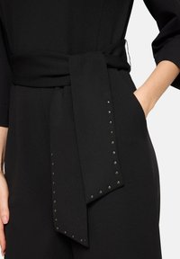Czarny kombinezon z szerokim paskiem z detalem węzła, ozdobiony metalicznymi ćwiekami wzdłuż kieszeni i brzegu. Gładka faktura.