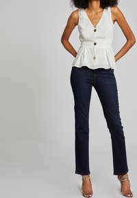 Bílý peplum top s hlubokým výstřihem do V a třemi zlatými knoflíky, spárovaný s tmavými rovnoběžnými džíny a sandály se zdobenými popruhy.