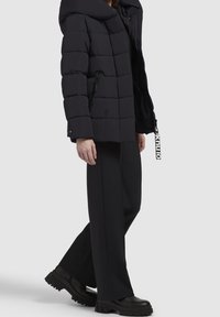 Henkilö, jolla on musta toppatakki hupulla, leveälahkeiset mustat housut ja paksupohjaiset mustat kengät, seisoo yksinkertaisen taustan edessä.