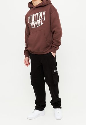 Ung person iført en brun hættetrøje med teksten "Multiply Apparel" og sorte cargobukser med logo-patch, kombineret med hvide sneakers.