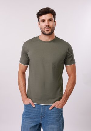 Homme barbu portant un t-shirt manches courtes vert olive et un jean bleu, debout les mains dans les poches devant un fond clair.