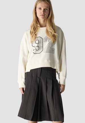 Blonde Frau trägt ein weißes Sweatshirt mit der Nummer 92 und einen schwarzen plissierten Rock mit Nadelstreifen, steht vor einem hellgrauen Hintergrund.
