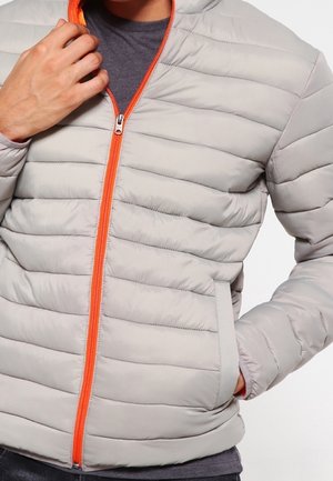 Homme portant une veste matelassée gris clair avec une fermeture éclair orange, une main touchant le col, l’autre main dans la poche latérale, chemise grise en dessous.