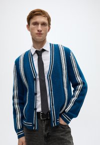 Μπλε πλεκτή ζακέτα με λευκές ρίγες, με V-λαιμόκοψη, κουμπιά μπροστά και υφή υφάσματος. Φορεμένη πάνω από λευκό πουκάμισο και γραβάτα.