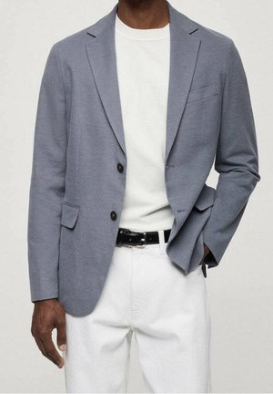 Homme portant un blazer bleu clair, un t-shirt blanc à col rond, un pantalon blanc et une ceinture noire, debout avec une main dans la poche.