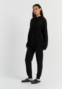 Felpa nera oversize con cappuccio e cordini, abbinata a pantaloni neri affusolati. Indossata con scarpe slip-on nere con un accento dorato.
