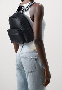 Mochila de cuero negro con forma redondeada, que cuenta con un bolsillo frontal y correas ajustables. La correa muestra el logo "HUGO".