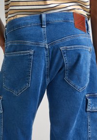Vista trasera de una persona que lleva pantalones cargo de mezclilla azules con una etiqueta de cuero marrón y una camisa de rayas beige.