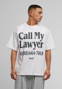 Hvit oversized t-skjorte laget av bomull med svart tekst som sier "Ring min advokat" og et telefonnummer, med korte ermer og rund hals.
