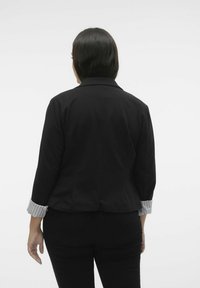 Sort blazer med trekvartærmer. Kontrasterende stribede skjorte manchetter er synlige. Glat stof, skræddersyet pasform og struktureret krave detajle.