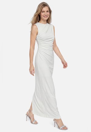 Vrouw in een witte mouwloze geplooide maxi-jurk, lopend, draagt zilveren hoge hakken sandalen, glimlachend, tegen een effen lichte achtergrond.