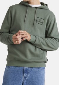 Green hoodie made of soft fabric with a kangaroo pocket, drawstrings, and a black rectangular logo patch on the chest. Worn with blue jeans.