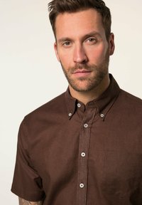 Brązowa koszula krótki rękaw z teksturowanej tkaniny, z kołnierzykiem typu button-down, kieszenią na piersi oraz białymi guzikami przy listwie.
