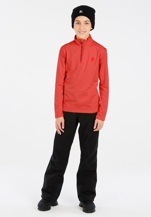 Jong persoon staat, draagt een rood shirt met lange mouwen en een kwartrits, zwarte broek, zwarte laarzen en een zwarte muts.