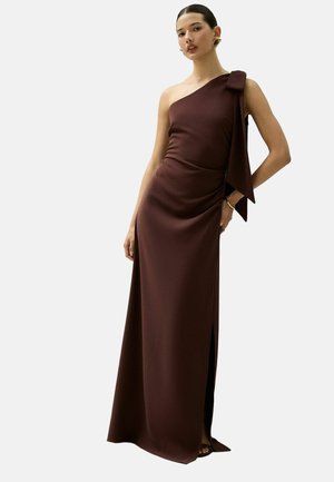 Frau trägt ein bodenlanges, dunkelbraunes Ein-Schulter-Kleid mit einer großen Schleife auf der Schulter und einem seitlichen Schlitz, steht vor einem schlichten Hintergrund.