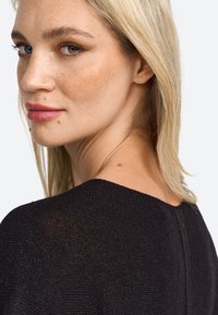 Czarny materiał o fakturze, luźny krój, z szerokim dekoltem. Noszony przez modelkę z długimi blond włosami i naturalnym makijażem.