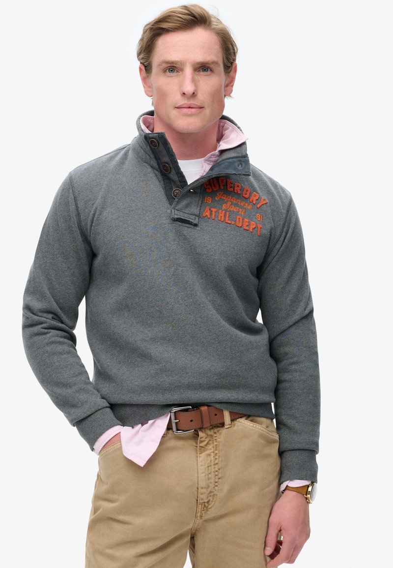 Pull-over gris avec col et détails à boutons, arborant un logo brodé rouge. Porté sur une chemise rose clair, associé à un pantalon beige.