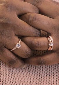 Due anelli sulle mani: uno è una sottile fascia d'oro con una pietra preziosa centrale; l'altro ha un design intrecciato più ampio con pietre trasparenti, entrambi in oro rosa.
